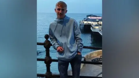 A young man wearing a blue zip up hoody is stood with an elbow leaning on a railing with the sea and a pier behind him.
