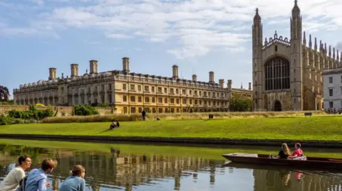Three students sit in the foreground by the river Cam, opposite Kings College Cambridge on a sunny day. Two more students are in a punt on the river passing by in front of them. 