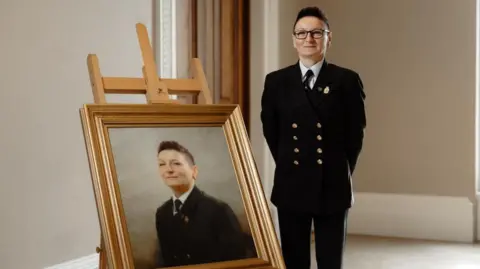 Karolina Jabryzk, wearing a formal dark uniform with brass buttons, stands beside an easel displaying a framed portrait of her, set against a neutral background. The setting is a room with light-coloured walls.