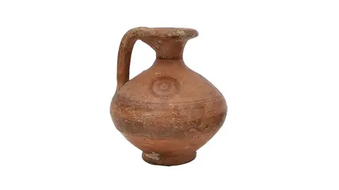 The Cotswold Auction House A small, reddy brown Cypriot jug, which dates back to 750 BC against a white background. The jug has a bulbous middle, a skinny neck, and a handle. There is a striped pattern around the middle with a swirl nearer the neck.