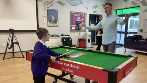 James Grant / BBC Boy in school jumper holds a snooker cue on a table while Kyren Wilson stands opposite with arms outstretched.