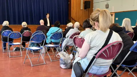 An image of the public meeting - we see Gordon McKee at the front of a community hall, addressing the local people who are sitting in fold-out chairs.