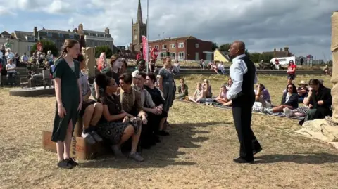 Richard Daniel/BBC Children taking part in an outdoor performance. They are wearing 1930s-style clothes and are watched by a small audience