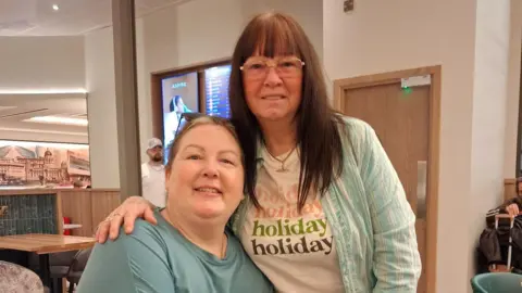 Rebecca is sat on the left of the photo and her mum Delia is stood to her right with her arm around her. Rebecca is wearing a blue t-shirt. Delia is wearing glasses and a stripy shirt over a white graphic t-shirt which says 'holiday' four times.