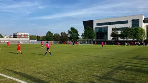 Two women's football teams, wearing red and pink kits, playing a match on an outdoor pitch. A black and white college building is in the background behind some trees.