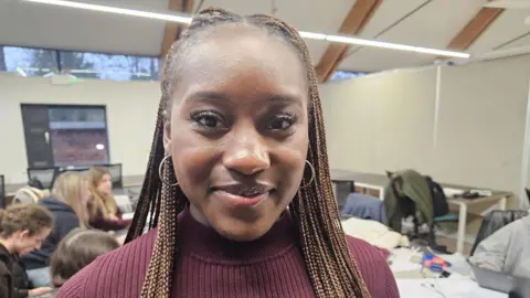 Mayah Baidoo has long dark brown braided hair, she is wearing hooped earrings and is smiling at the camera. She is wearing a burgundy high necked jumper. In the back ground other students can be seen working at desks.