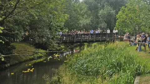 University of Buckingham People gathered on a river bank and on a pedestrian bridge over the river watching a rubber duck race. There are trees on either side of the river.