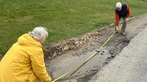 Two people holding a tape measure over a large pothole on the side of the road