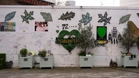 PA Media The memorial beneath Grenfell Tower, with a green heart with the words Grenfell written in the middle, the number 72 above it, alongside the words 'WHY', some pictures and large posters of leaves with the wording 'Justice'.