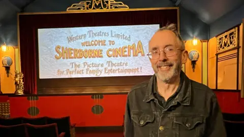 Mark Cunningham, who has silver glasses and a beard, and is wearing a black denim jacket, smiles at the camera from inside the small cinema's auditorium. Behind him is the main screen welcoming people to the cinema. The screen has deep red velvet curtains around it, and on either side are orange painted walls with traditional 1950s style lamps. There is a small ornate golden statue to one side of the screen.