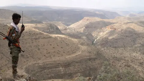 BBC/Amensisa Ifa A young man in camouflage clothing stands on the left of the picture with a gun slung over his shoulder. He is holding a walkie-talkie and is looking out over a valley amid the vast expanse of barren mountains. 