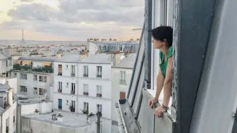 Kate Abby Kate bersandar ke luar jendela saat dia memandangi cakrawala kota. Menara Eiffel dapat dilihat di latar belakang sebelah kiri.