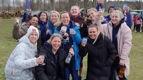 Sunderland Strollers A group of 10 women and one man smiling into the camera. Two of them are holding bottles of prosecco and the others are holding up plastic glasses. They are wearing coats and hats.