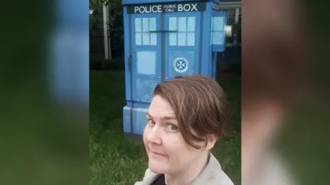 Erica Moore Erica with short dark hair, wearing a black t shirt and beige blazer, stood in front of the Tardis