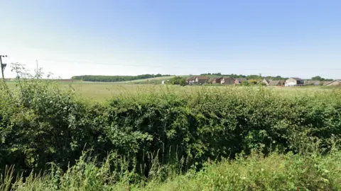 Google A small hedge is in front of a large field which goes across the whole picture. On the right there is a small housing estate. 