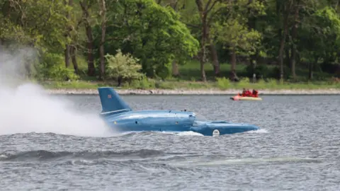 Matthew Willis A plume of spray rises from the back of Bluebird as it travels across Coniston Water. The machine looks like a cross between a boat and a plane. Painted blue, it has a tail fin. Trees can be seen on the shoreline.