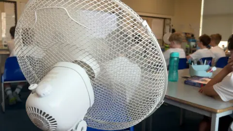 Generic shot of a white electric fan in a school classroom, with pupils in the background sitting at desks
