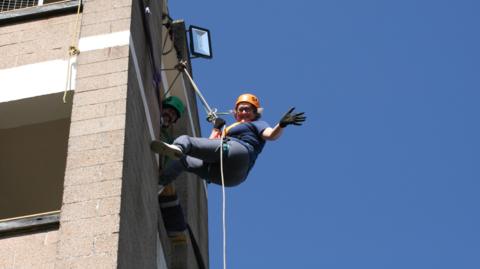 Sarah Spencer at the start of her abseil, waving to the camera and smiling against a clear blue sky. 