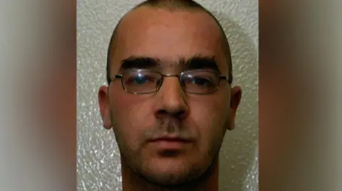 A man in his 20s, with close-cropped dark hair, thick eyebrows and metal-rimmed glasses, looking at the camera.