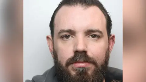 A police mugshot of Matthew Whatron, who has black hair, a beard and lip, ear and nose piercings.