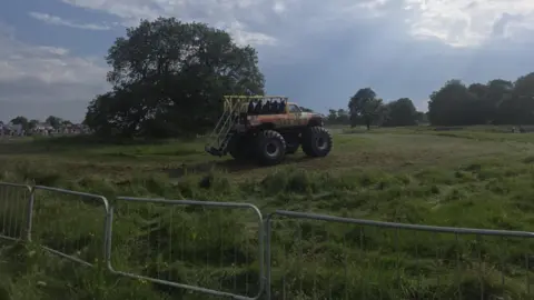 Claire Southern People are sitting on an orange monster truck that is in a field. There is a metal fence cordoning off the field.