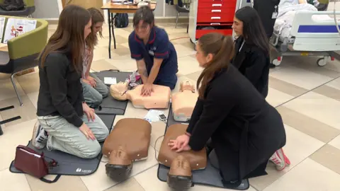 Richard Knights/BBC A group of five women kneel on mats around mannequins  used for CPR training, one is a nurse in uniform. Behind them is a hospital trolley