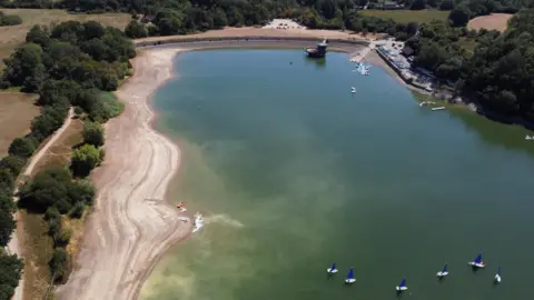 Reuters Ardingly Reservoir with small boats and kayaks on the water, seen from above. The water level has dropped and a shoreline is visible.