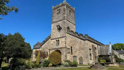 St Mary's Church has a large central tower, roughly three storeys tall. It has a crenelated roof sloping down from the middle of the tower. A small monument stands on the grass outside the church, there are three steps with a small pillar in the centre. The surroundings of the church are lush and green with bushes and trees surrounding it.