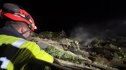 Kevin Adams Man in high-vis jacket and helmet at the top of cliff edge looking down at two other men in high-vis and hard hats abseiling down cliff.