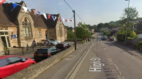 Google Box Primary School building to the left with a road on the right and zebra crossing in the distance with a group of people about to cross. There is bunting hanging down the road.