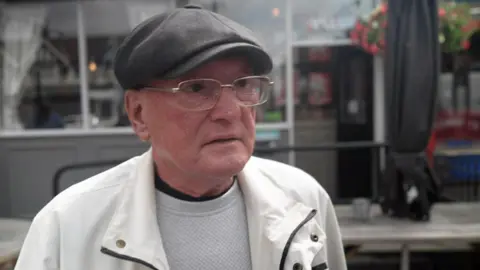 Paul is wearing a dark grey flat-cap on his head, rectangular metal glasses on his face, a white jacket over a grey top. He is standing in front of a grey pub building with floral hanging baskets.