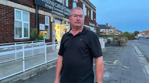 A man in glasses and a black polo shirt. He is standing in front of a cafe which is in a red brick building with white railings in front of it. The man has short grey hair and he does not look happy. A street can be seen behind him with houses and parked cars.