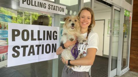 Josie with her pooch Arthur at the polling station in Henley, Oxfordshire. Josie is wearing a white t-shirt with mushrooms printed on it and grey trousers. She has her hair in a pony tail and she is holding her dog Arthur. They are stood outside a glass fronted building which has a sign reading "polling station" in the window
