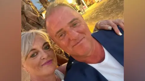 Melanie Graham A "selfie" picture of McDonald and Mel on a beach. She has short blonde hair and is wearing pink lipstick. She has her arm around him and he is wearing a white t-shirt and blue suit waistcoat. They're both smiling.