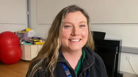 Sinéad is smiling at the camera and has long blonde hair. She is wearing a black fleece with a green polo top underneath.