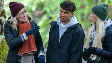 Three members of series three of the BBC's UK version of The Traitors stand talking to each other intensely. 