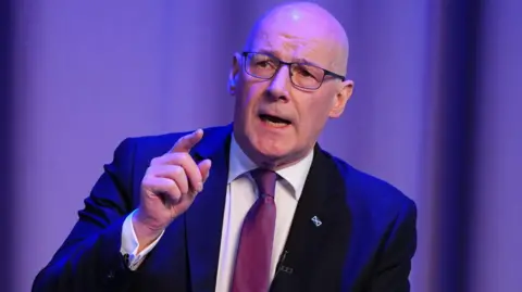 John Swinney, who is bald and is wearing glasses, is wearing a blue suit, white shirt and purple tie and is sitting in front of a purple background. He has a saltire tie-pin on his left lapel.