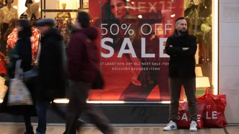 Reuters Shoppers walking past a Next store with a red sign in the window saying 