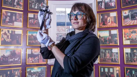 Charlotte Graham Dr Hannah Thomas holds up a small wooden cross with the figure of Jesus on it in gloved hands. Dr Thomas wears a grey jumper, dark grey scarf and blue-rimmed glasses. She has short brown hair.