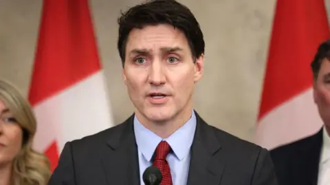 Justin Trudeau pictured at a press conference speaking into a microphone. There are two ministers and Canadian flags visible in the background. 