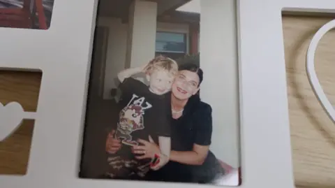 Geraldine, wearing a black top and white necklace, hugs her son from behind. He has blond curly hair and is wearing a black t-shirt with a cartoon on the front.