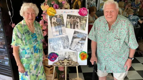 Mary and Eric stand on either side of a large display board mounted on an ornate easel. The display board contains several vintage wedding photographs and is decorated with colorful paper flowers. At the top of the board, the visible text reads "11 August 1951", suggesting the date of a wedding.
