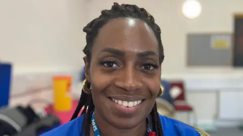 BBC Makini Jones is wearing an NHS lanyard and has dreadlocked hair, tied back, and is wearing looped gold earrings 