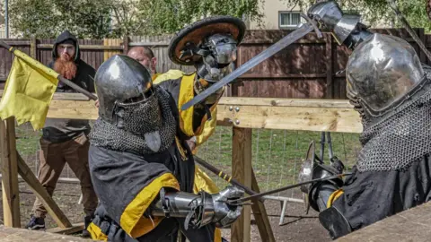 Knight Illuminations Close-up action scene of armoured combat inside a wooden-fenced arena. Two people in steel helmets and chainmail engage with swords at close range, blades crossed. One raises a sword overhead while the other blocks. A referee or observer stands behind holding a yellow flag, with trees and a residential building visible in the background.