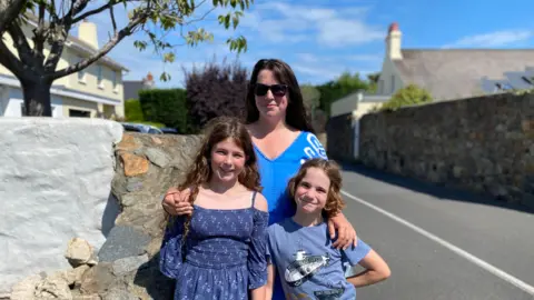 BBC Claire with her two children stood on a pavement. A road is on the right. The woman has sunglasses on and dark hair. They are all smiling towards the camera.