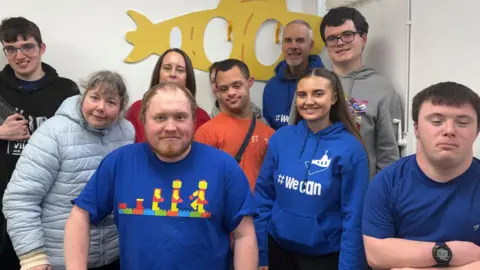 Ten people stand in the corner of a white room with a cut out of a yellow submarine behind them. From left to right, stands a young man who is a member of the group wearing a black hoodie, holding a black tablet which helps him communicate. He has dark brown hair and wears glasses. Next is a woman with grey hair who wears a pale blue puffer coat. The lady next to her has dark brown hair with a red t-shirt on, she works for the charity. There is then two young men, one wearing a grey hoodie and the other an orange t-shirt. There is then another member of the charity. He stands in the back corner and has grey hair. He wears a staff hoodie with the words 'we can' on. There is then a tall young man wearing a grey hoodie and black framed glasses. At the front of the picture is a young woman and two young men- all wearing navy blue and smiling at the camera.