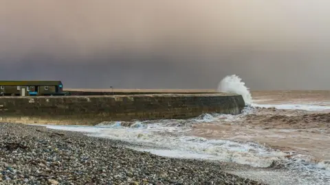 Ian Capper View from the shingle beach of the seaward side of the Cobb breakwater. It is a stormy-looking sky and the sea is crashing against the wall. On the left on top of the wall are two buildings with pitched roofs and chimneys.