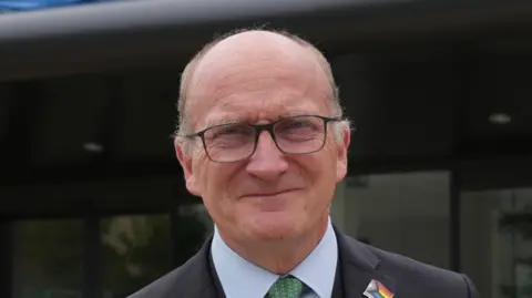 Nikki Fox/BBC Nick Hulme smiles at the camera. He is balding with some thin grey hair around the edge of his head. He wears glasses and a black suit with a white suit and green tie. A Pride flag badge is pinned to his collar.