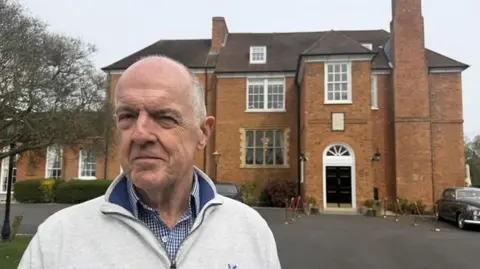 A man wearing a blue checked shirt with a pale grey jumper over the top stands at the forefront of the picture, looking off to the left of the camera. Behind him is a red brick building, with tall windows and a black door.