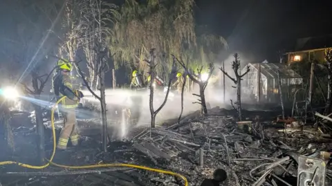 Firefighters hose down a burnt and destroyed garden. Trees can be seen burnt with rubble strewn across the garden. 